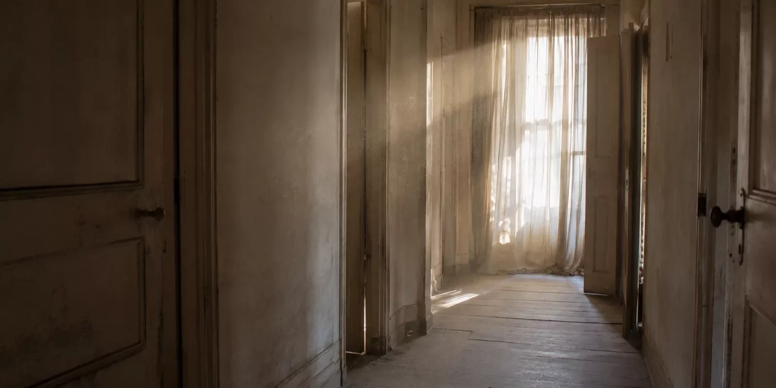 Couloir d'une maison ancienne inoccupée, poussière en suspension dans la lumière filtrée par un rideau