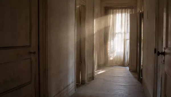 Couloir d'une maison ancienne inoccupée, poussière en suspension dans la lumière filtrée par un rideau